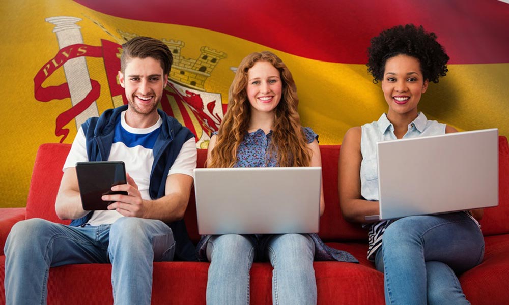 یک دختر مو فردار کوتاه مشکی کنار دختری مو طلایی با لپ تاپ های سفید روی مبل قرمز کنار وسری با یک تبلت مشکی نشسته و در حال لبخند هستند و پرچم اسپانیا بک گراند آن هاست - اقامت دائم اسپانیا از طریق تحصیل 