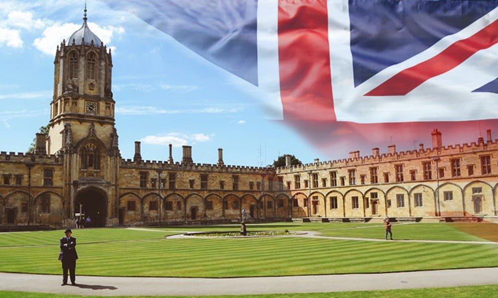 نمای فضای بیرونی دانشگاه کمبریج مردی مقابل ساختمان جلوتر از چمن ها ایستاده پرچم انگلیس هم به صورت فلو شده در آسمان دانشگاه به چشم می خورد - شرایط سنی تحصیل در انگلستان